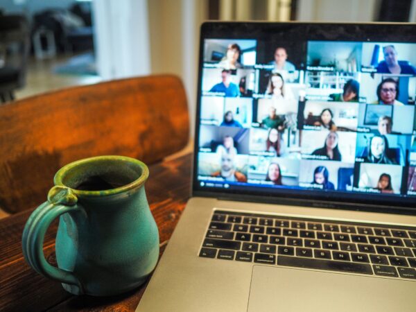 Die Fotografie zeigt einen aufgeklappten Laptop und daneben eine Tasse. Auf dem Laptop sind eine große Menge Teilnehmende einer Videokonferenz zu sehen.