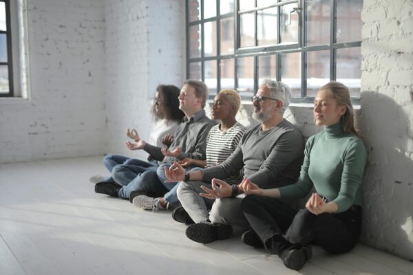 Die Fotografie zeigt eine Gruppe Menschen verschiedener Phänotypen angelehnt an eine Wand und im Schneidersitz bei einer Meditation