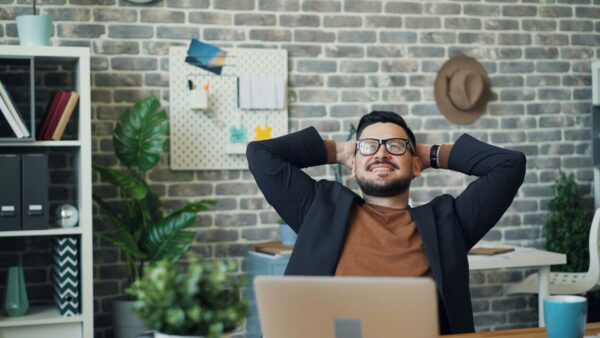 Das Foto zeigt einen junden Mann mit Brille hinter seinem Laptop am Schreibtisch sitzend. Er lehnt sich zurück mit den Händen hinter dem Kopf verschränkt und lächelt.